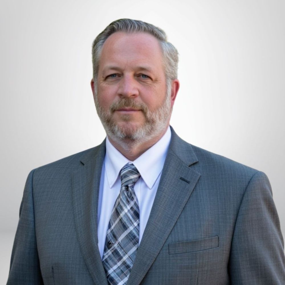Man in a gray suit with a plaid tie, beard, and short hair, standing against a light background.