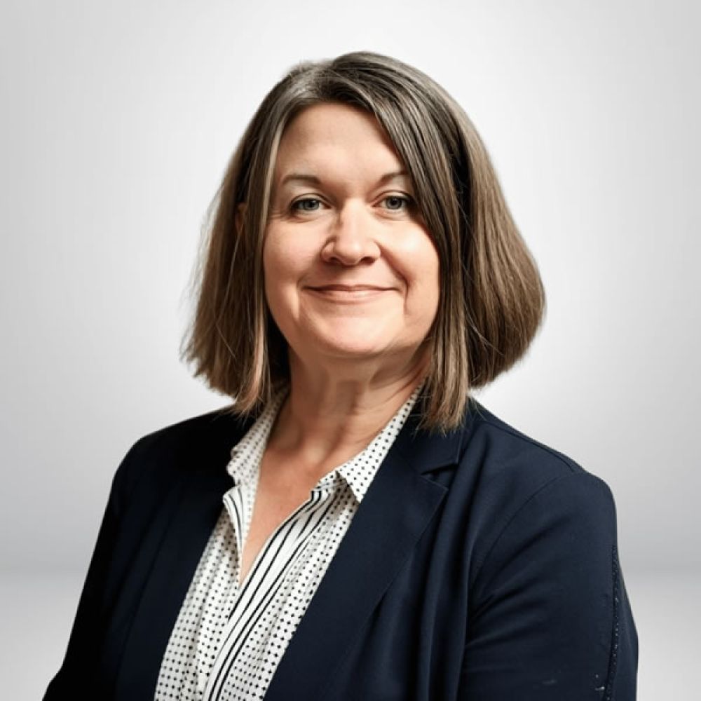 Professional woman smiling in a blazer, standing against a light background.