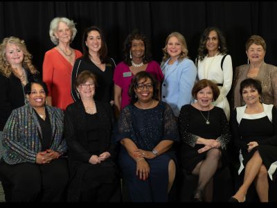 Group of women posing together at an event, wearing formal attire, smiling and standing against a black backdrop.
