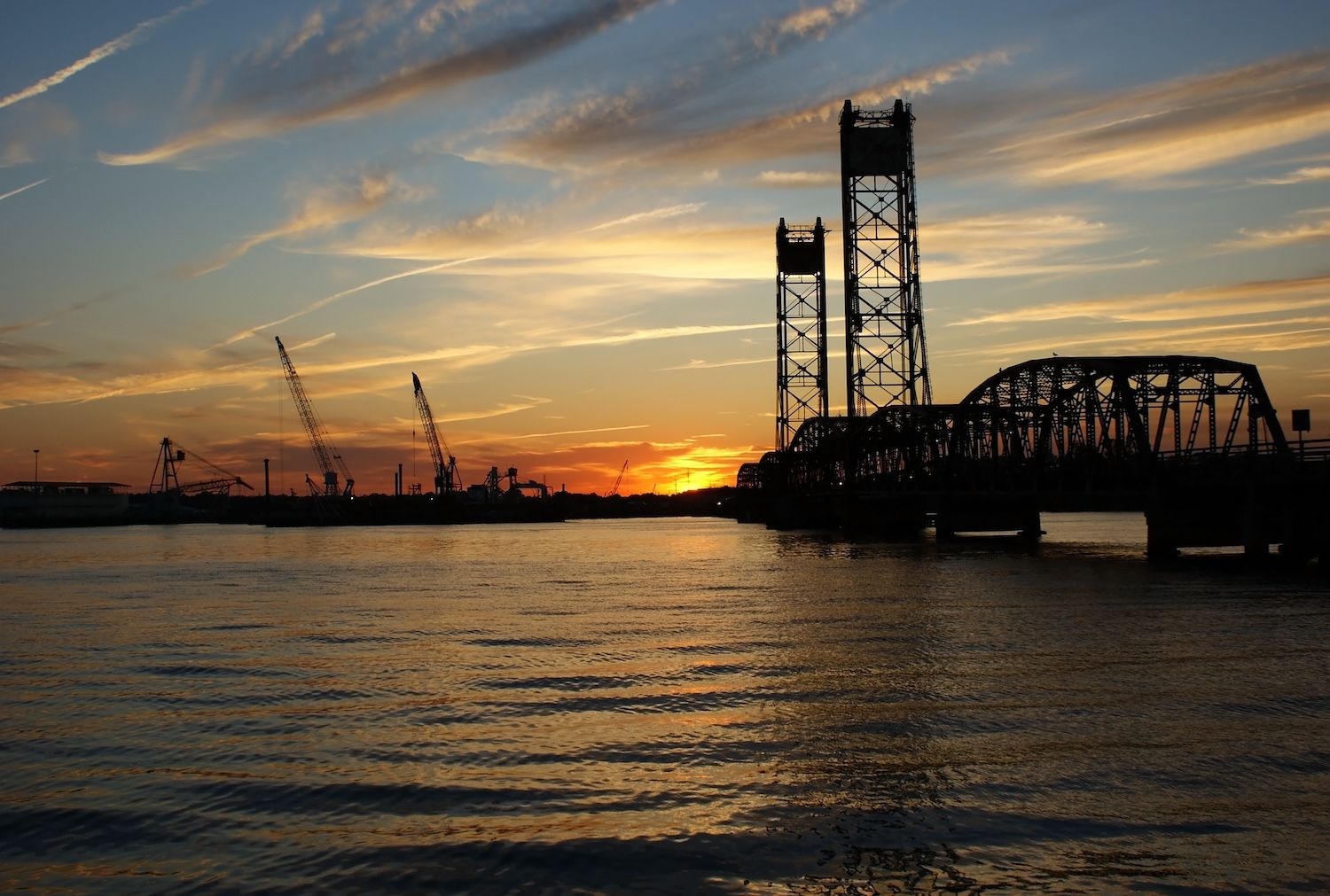Sunset behind a silhouette of a lift bridge and cranes over calm water, with vibrant skies filled with wispy clouds.