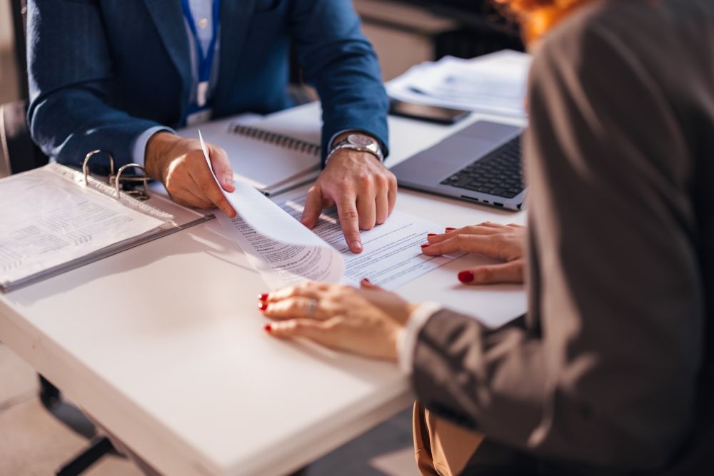 Business meeting with documents on table, two people discussing and reviewing paperwork in office setting.