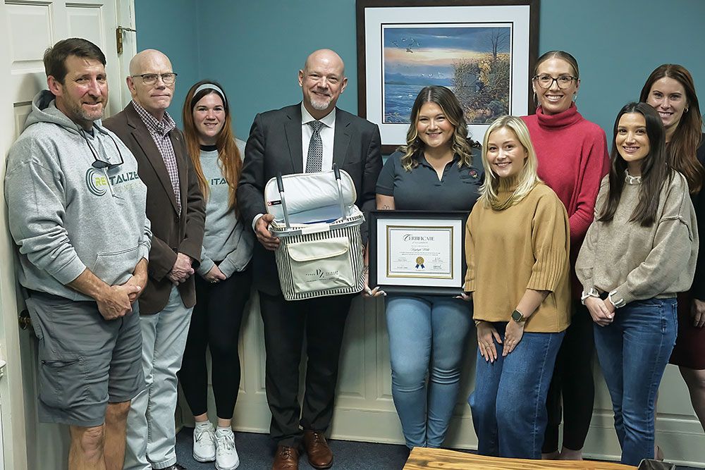 Group of people smiling indoors, holding a certificate and gift basket, celebrating an achievement in an office setting.