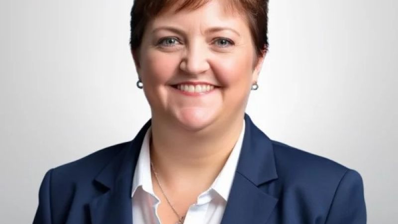 A professional woman in a navy blazer smiles in a studio portrait with a plain background.