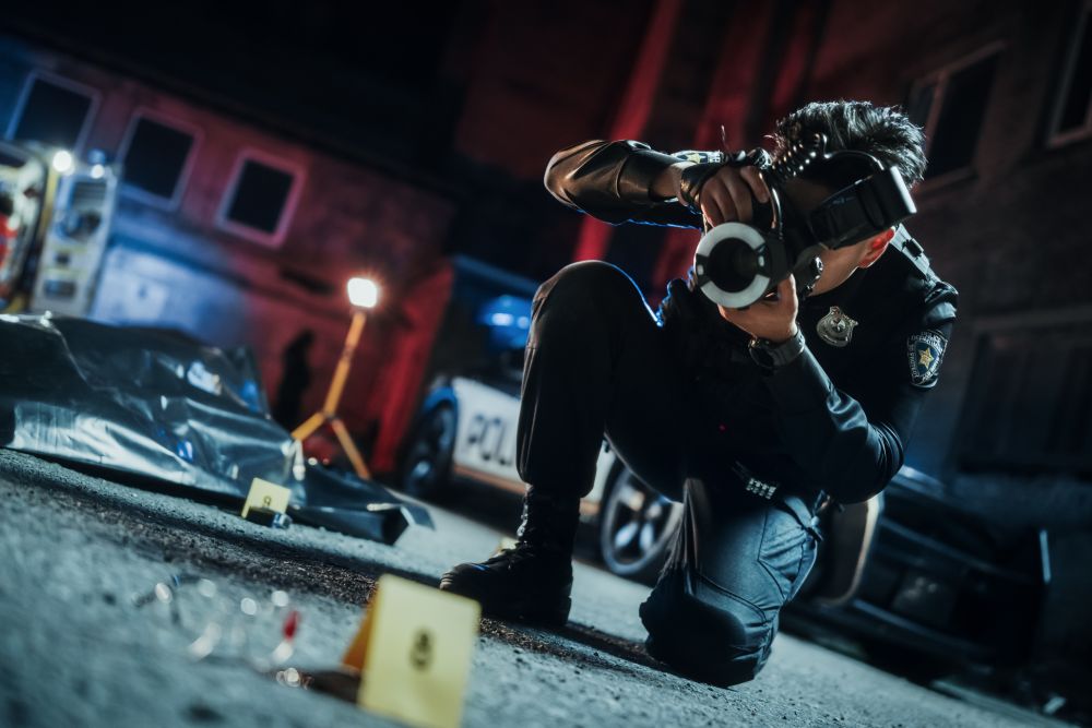 Tilted Shot: Asian Policeman Taking Pictures of Marked Evidence, the Victim's Bagged Corpse in the Background. Police Officer Documenting Bloody Glasses on the Floor For Further Investigation