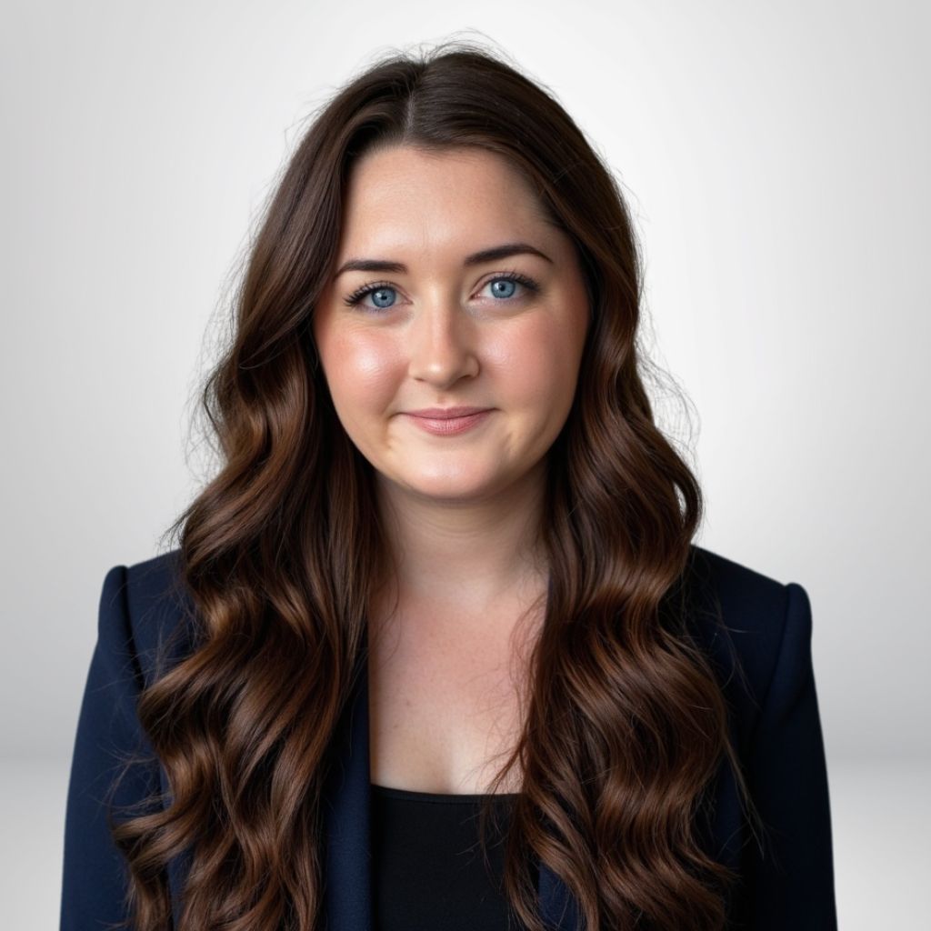 Portrait of a woman with long brown hair in a navy blazer, smiling softly with a neutral background.