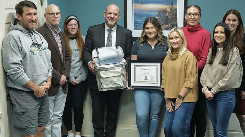 Group of people smiling indoors, holding a certificate and gift basket, celebrating an achievement in an office setting.