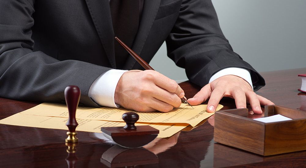 Businessperson signing document with pen at desk, surrounded by stamps and envelope.