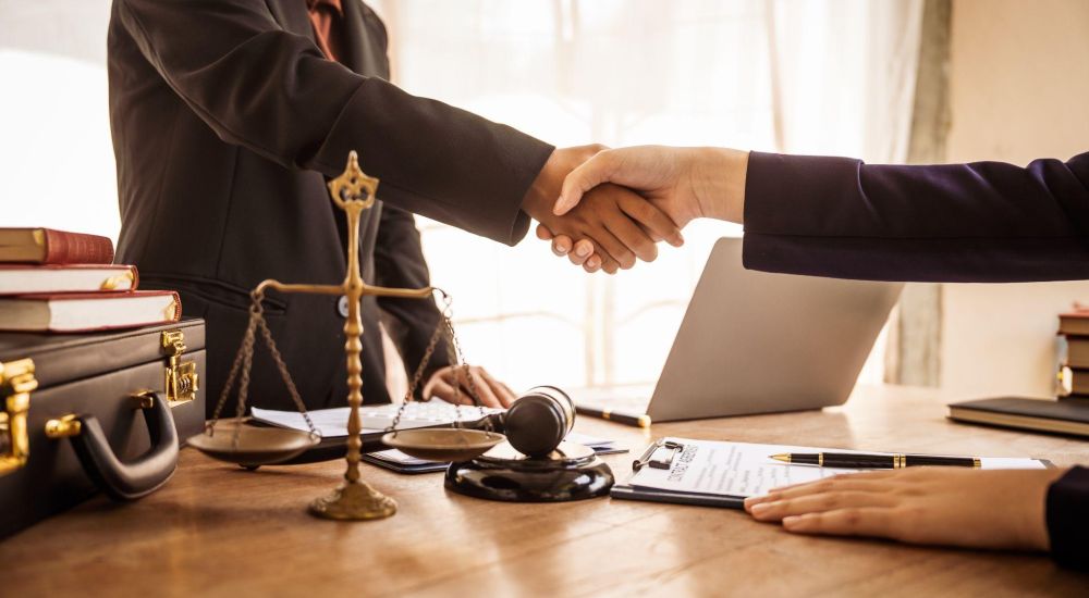 Two business professionals shaking hands over legal documents and scales of justice on a desk.