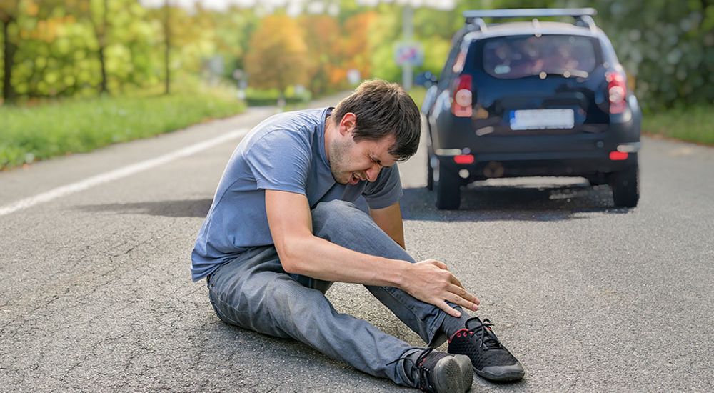 Injured man sitting on road after car accident, holding knee in pain, black car in background.