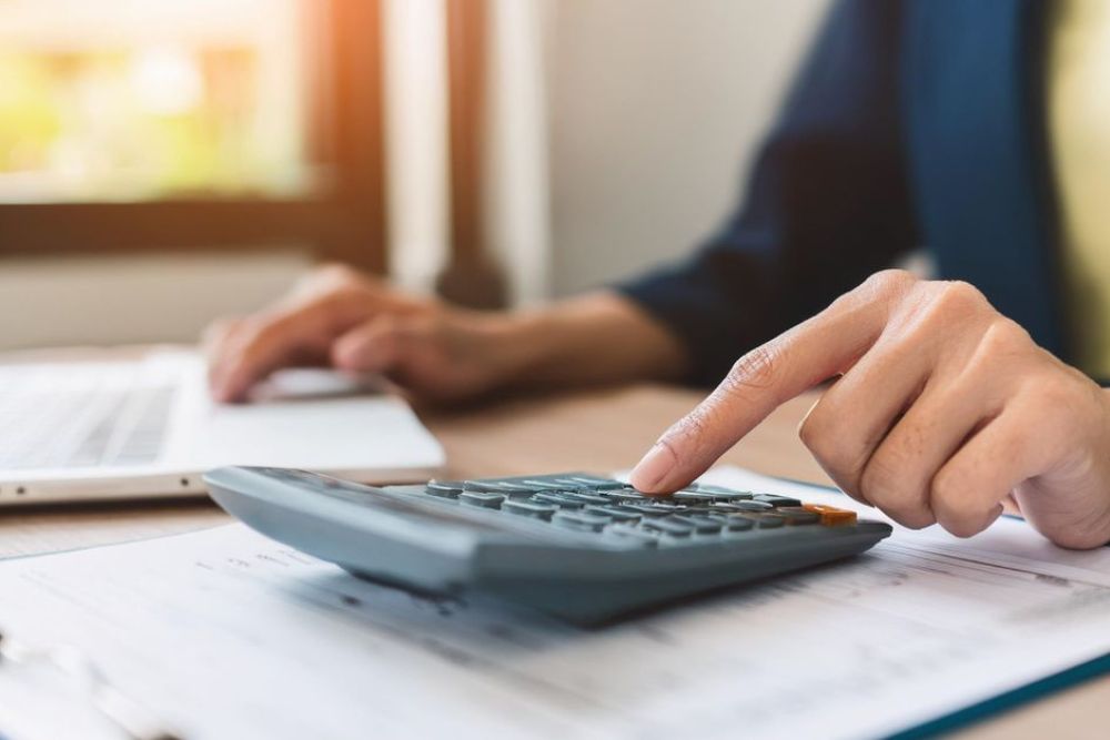 Person working on a laptop using a calculator, focused on financial calculations in a bright office setting.