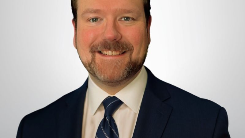 A smiling man in a suit and tie against a plain background.