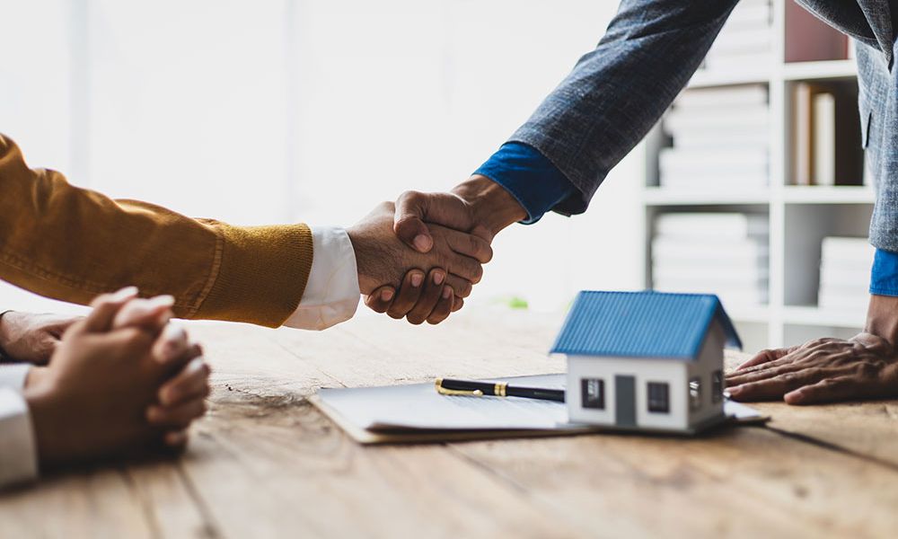 Handshake over a real estate contract with a model house, signifying a property deal.