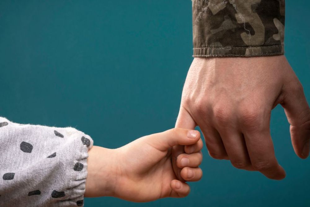 Child holding military parent's hand, symbolizing love and support against a teal background.