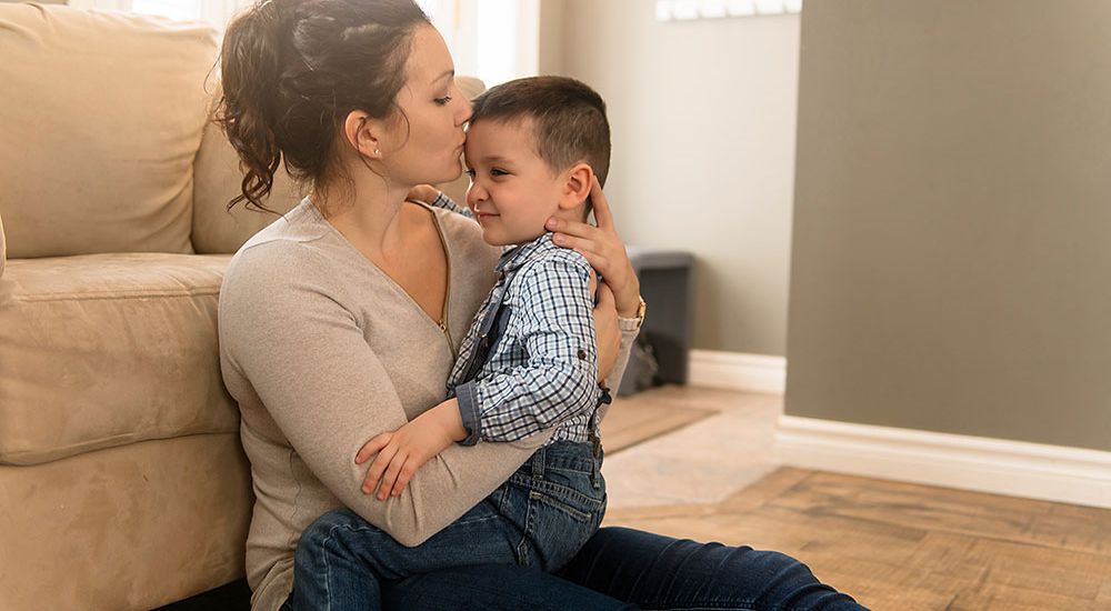 Mother lovingly kissing her young son on the cheek while sitting together on the floor at home.