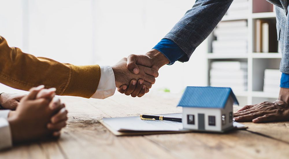 Handshake over a real estate contract with a model house, signifying a property deal.