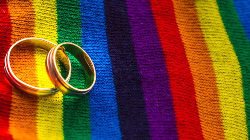 Two wedding rings on a vibrant rainbow-striped fabric, symbolizing LGBTQ+ pride and unity.
