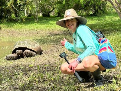 Woman poses with giant tortoise in lush, green forest, wearing hat and colorful backpack.