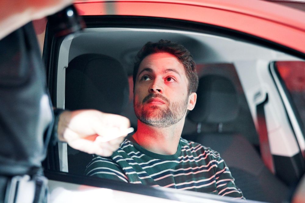 Driver in striped shirt stopped by police officer, exchanging documents through car window.