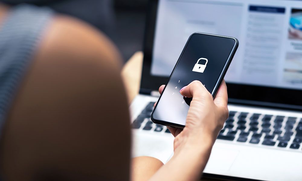 Person unlocking smartphone with security icon, seated at a laptop, illustrating cybersecurity and digital access.