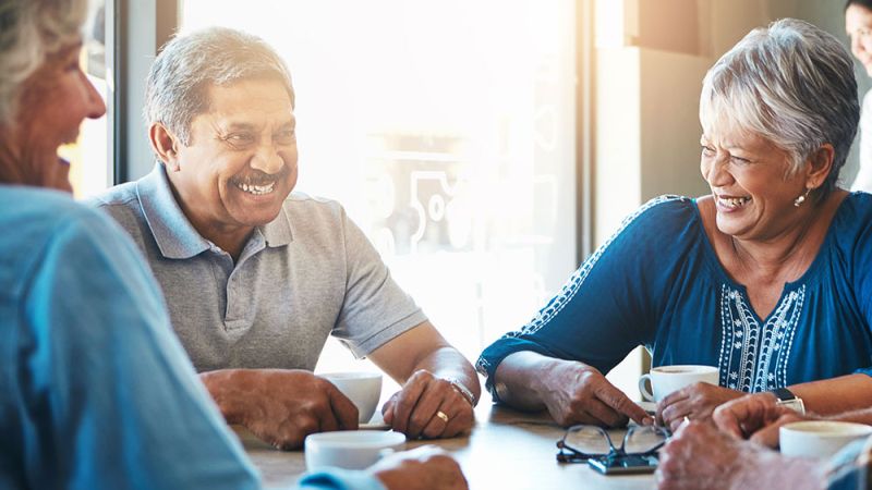 Senior friends laughing and enjoying coffee together at a cafe, sunlight streaming in through the window.