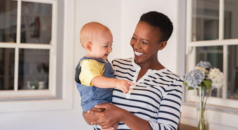 Smiling woman in striped shirt holds a laughing baby in a bright room with flowers in the background.