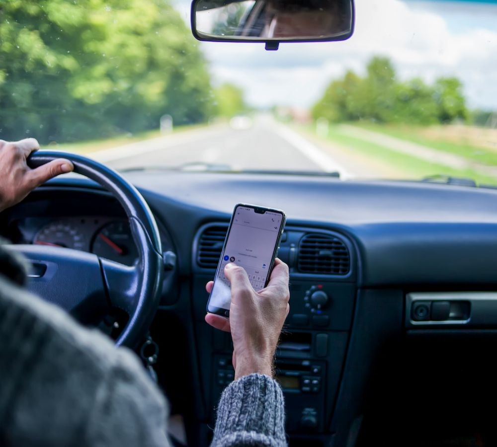 Driver using a smartphone while steering a car on an open road, illustrating distracted driving and safety concerns.