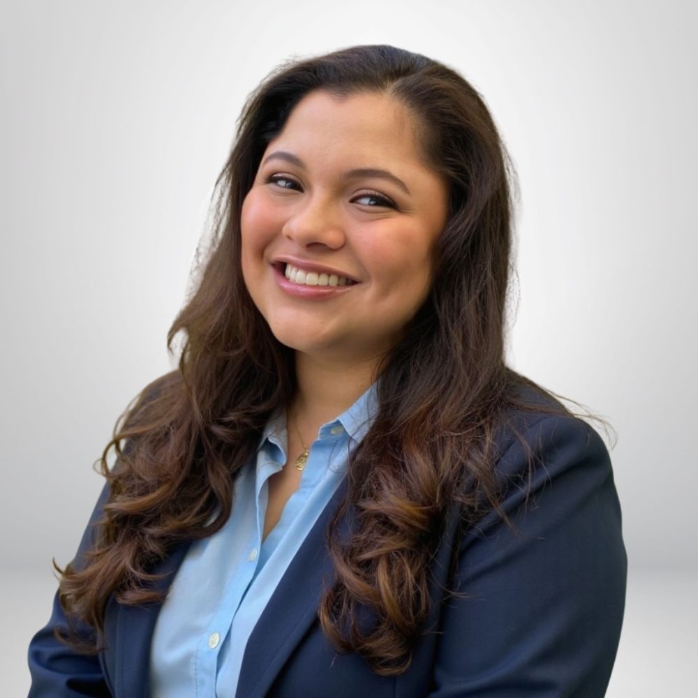 Smiling woman in a blue shirt and blazer, with long hair, posing against a simple background.