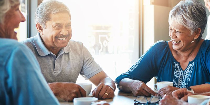 Senior friends laughing and enjoying coffee together at a cafe, sunlight streaming in through the window.