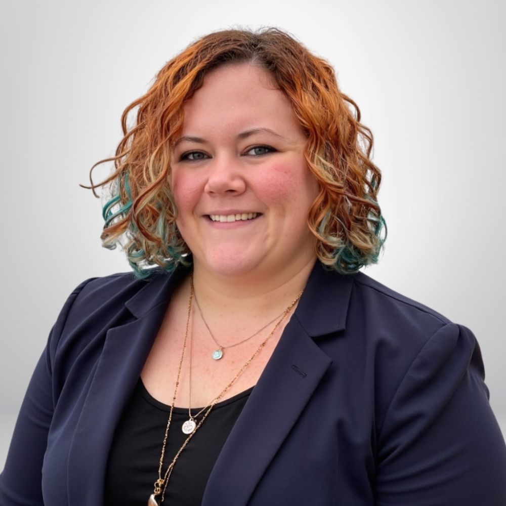 Smiling person with curly hair, wearing a navy blazer and layered necklaces against a plain background.