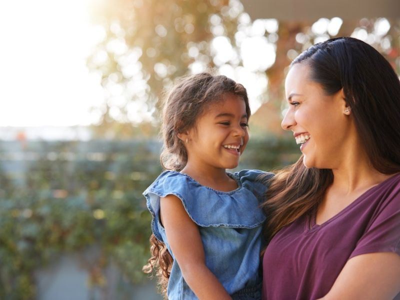Mother and daughter smiling at each other outdoors, enjoying a sunny day together.