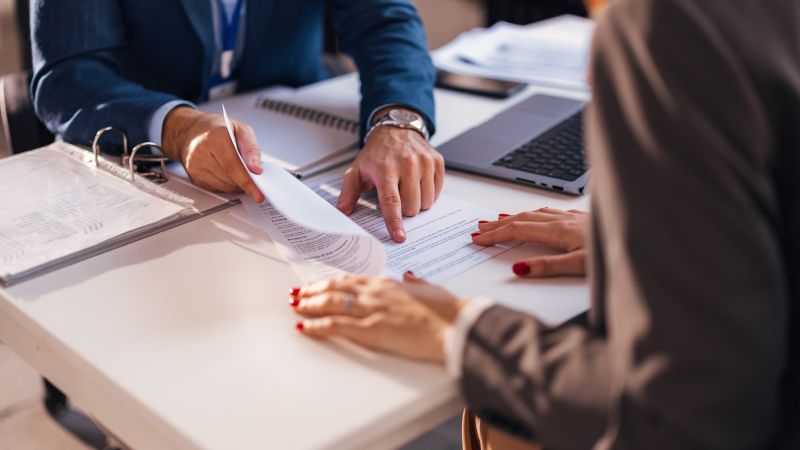 Business meeting with documents on table, two people discussing and reviewing paperwork in office setting.