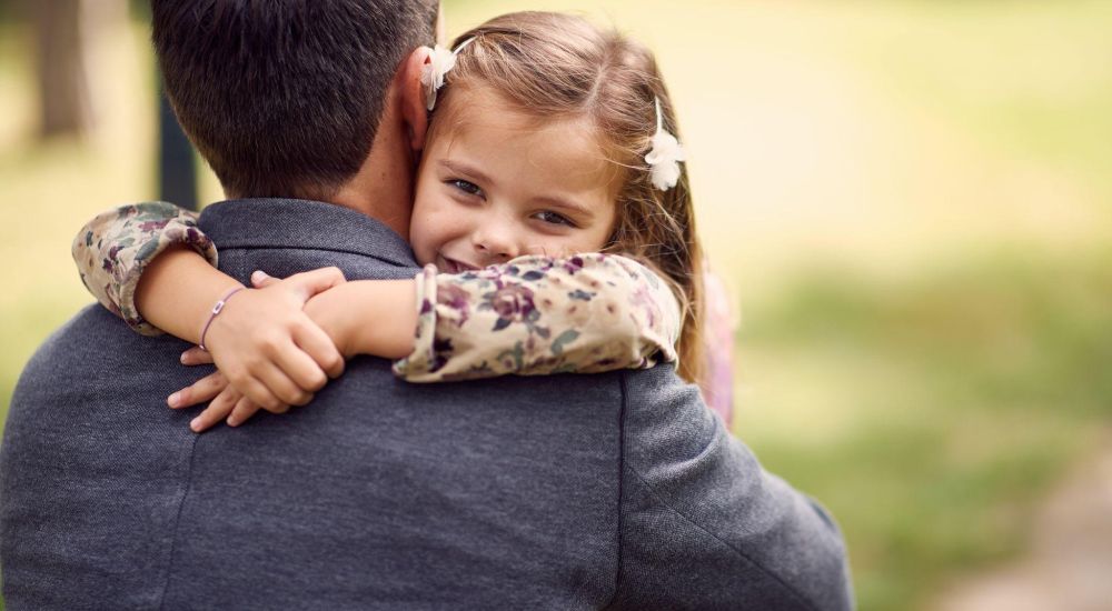 Father in gray suit hugging smiling daughter outdoors, highlighting family love and bonding.