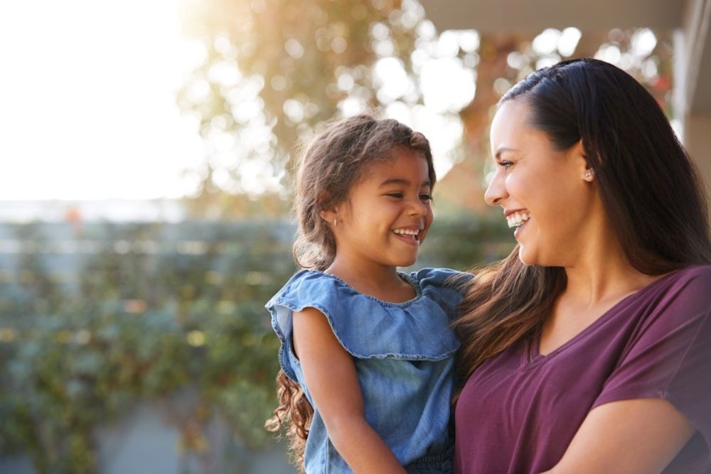 Mother and daughter smiling at each other outdoors, enjoying a sunny day together.