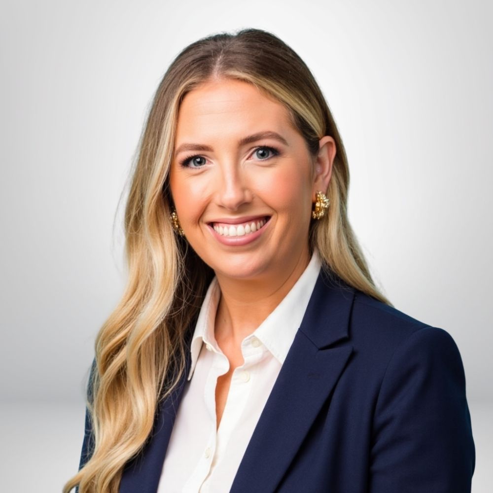 Smiling woman in a navy blazer and white shirt, with long blonde hair and gold earrings against a light background.