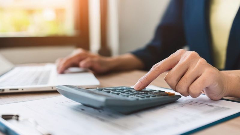 Person working on a laptop using a calculator, focused on financial calculations in a bright office setting.