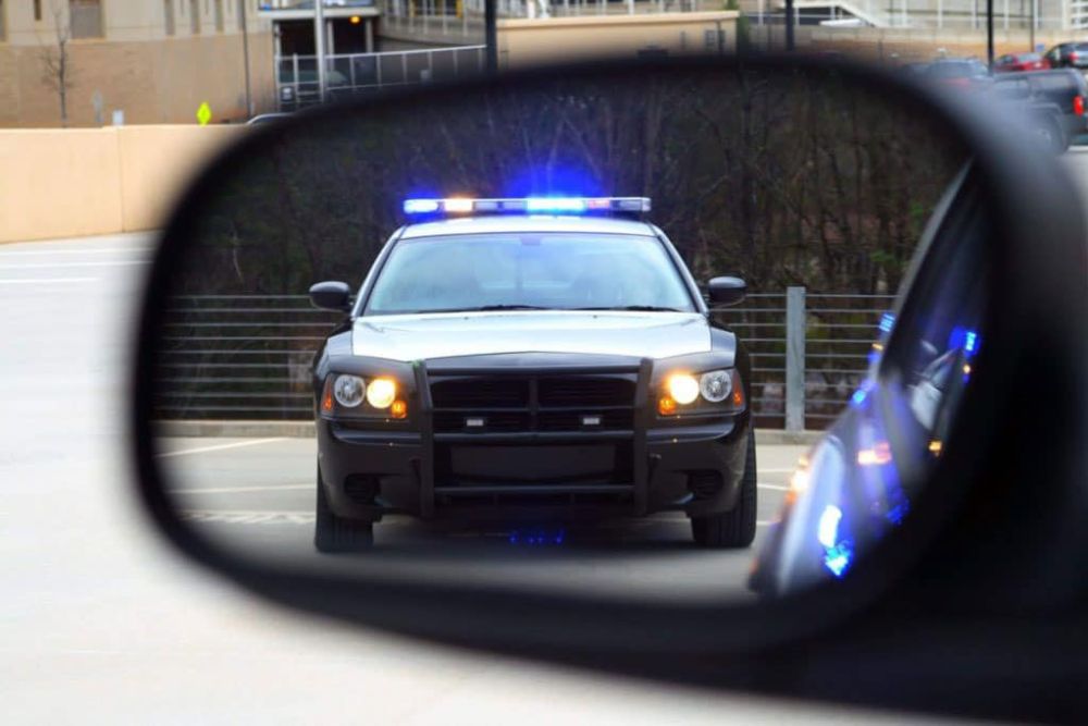 Police car with flashing lights seen in rear-view mirror on a road.