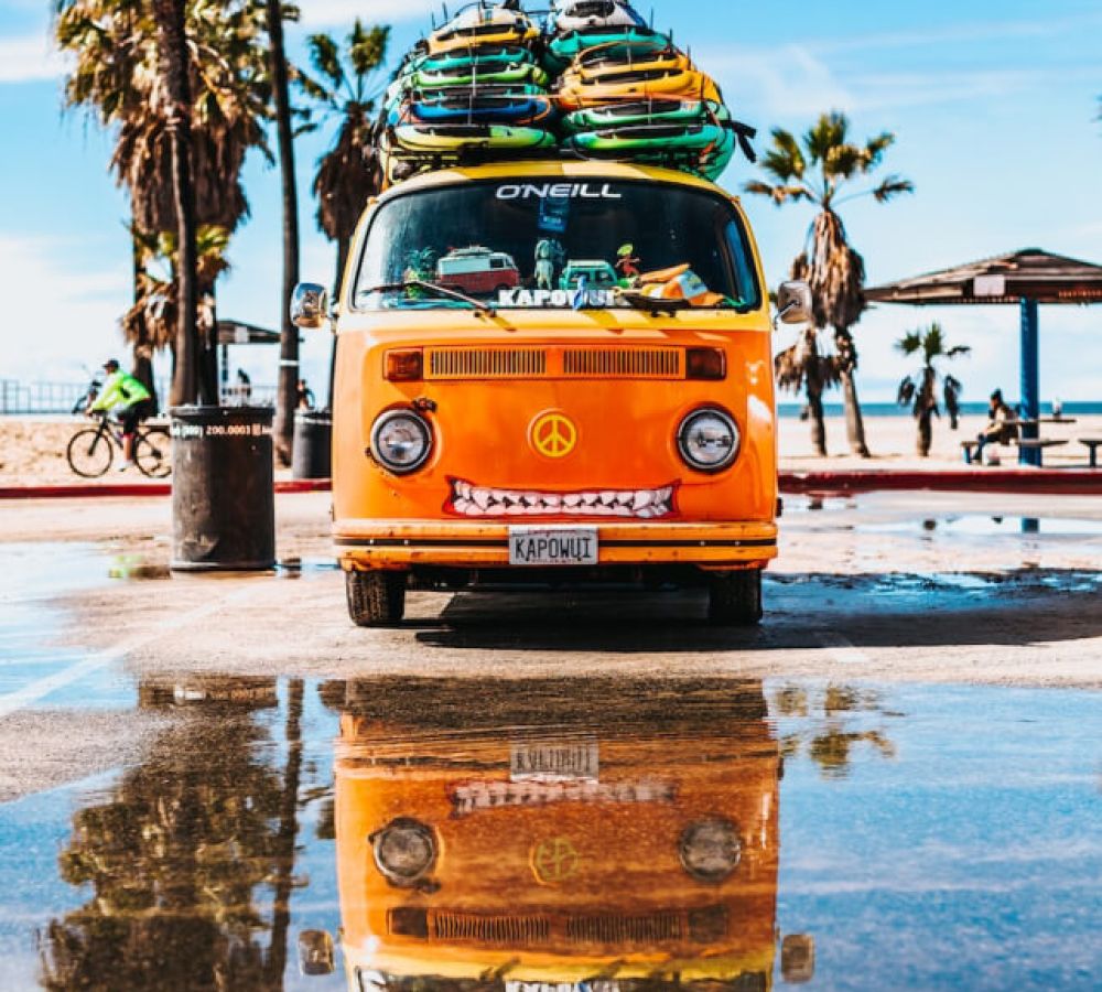 Orange vintage van with surfboards on top, reflecting in a puddle at a sunny beach scene with palm trees.