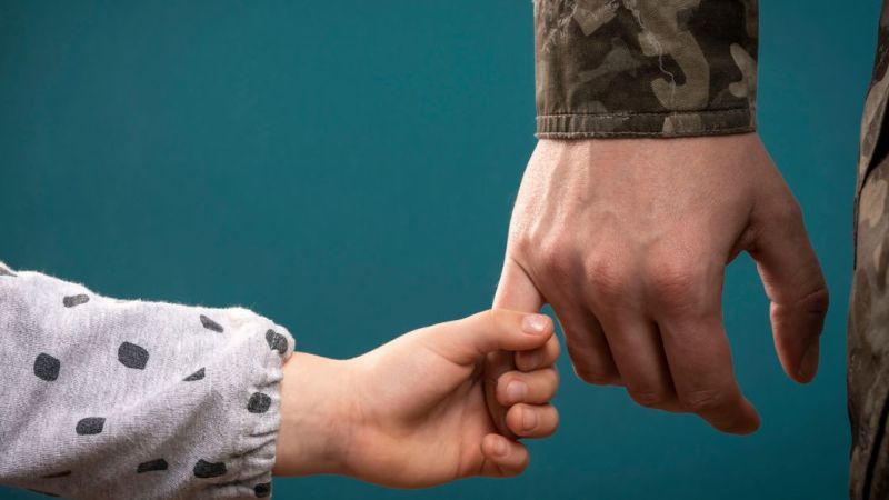 Child holding military parent's hand, symbolizing love and support against a teal background.