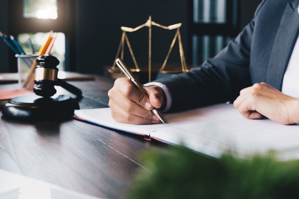 Lawyer writing at desk with gavel and scales of justice in background, symbolizing legal profession and justice.