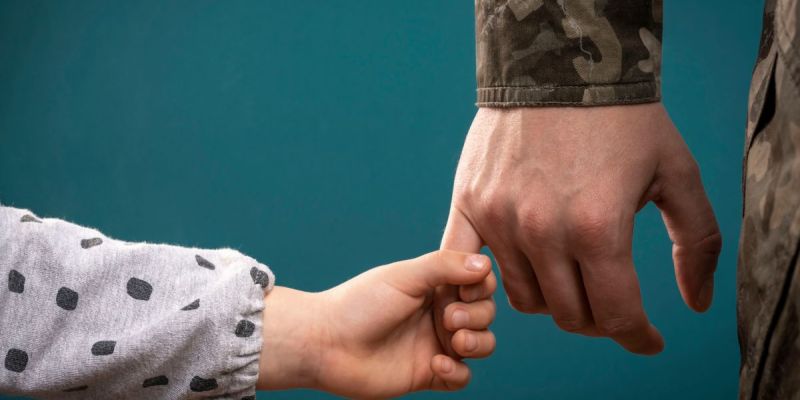 Child holding military parent's hand, symbolizing love and support against a teal background.