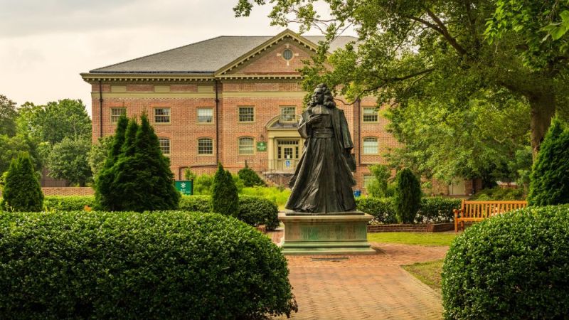 Statue in a lush garden, brick building in background, historical ambiance, sunny day, serene college campus setting.