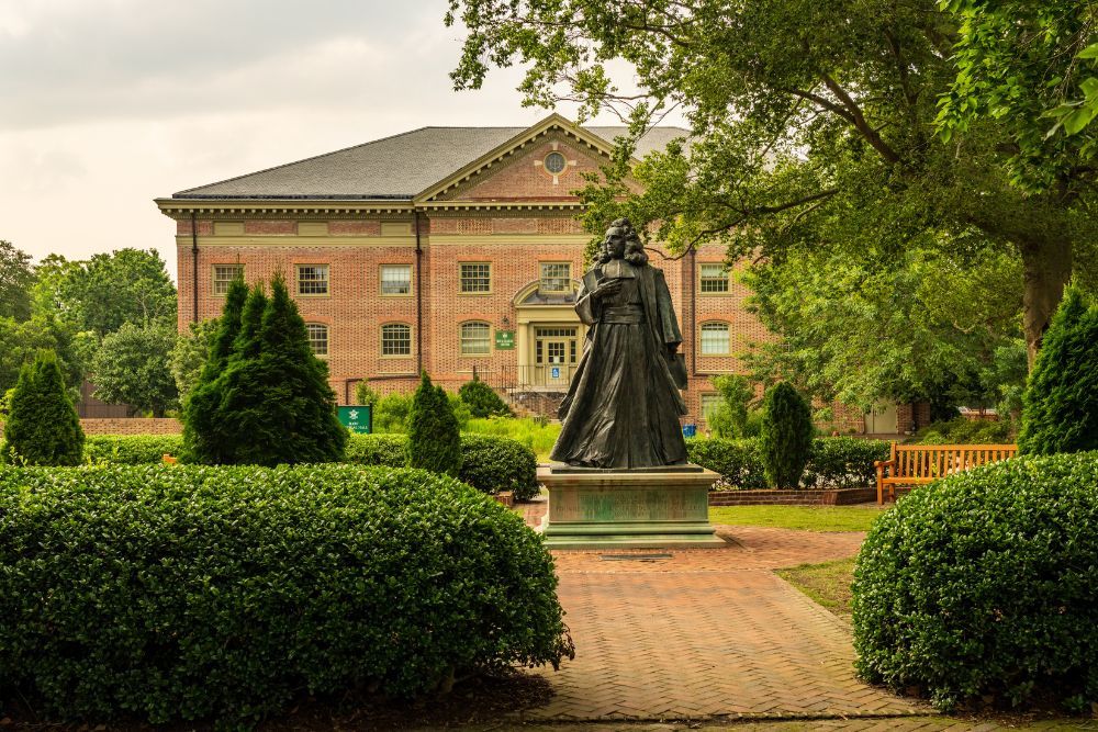 Statue in a lush garden, brick building in background, historical ambiance, sunny day, serene college campus setting.