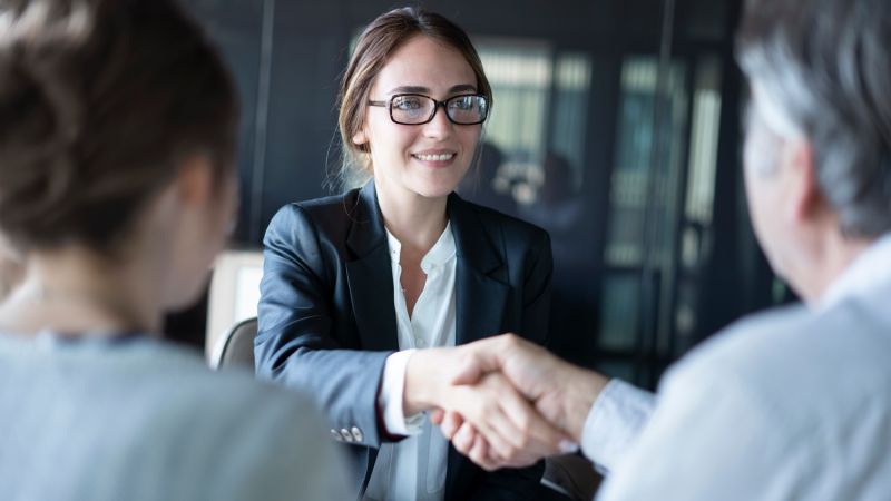 Businesswoman in glasses smiling and greeting colleagues with a handshake during a meeting, symbolizing successful negotiation.