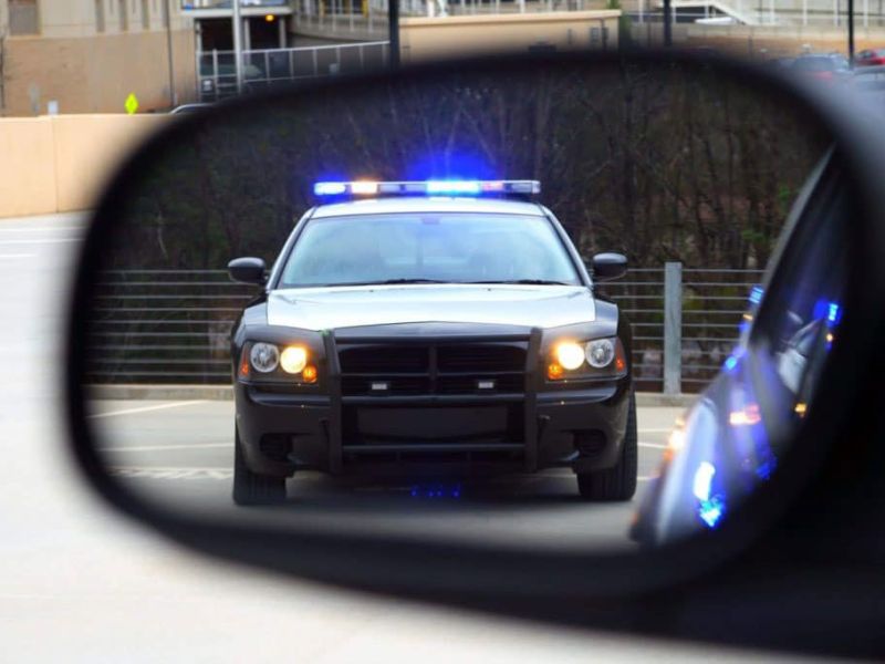 Police car with flashing lights seen in rear-view mirror on a road.