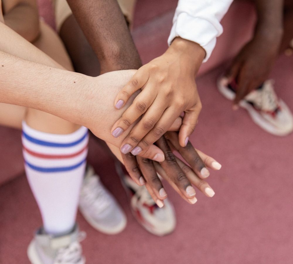 Diverse hands stacked together on a pink surface, symbolizing unity and teamwork, with striped socks and sneakers visible.