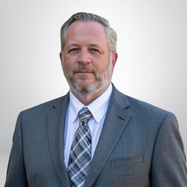 Man in a gray suit with a plaid tie, beard, and short hair, standing against a light background.