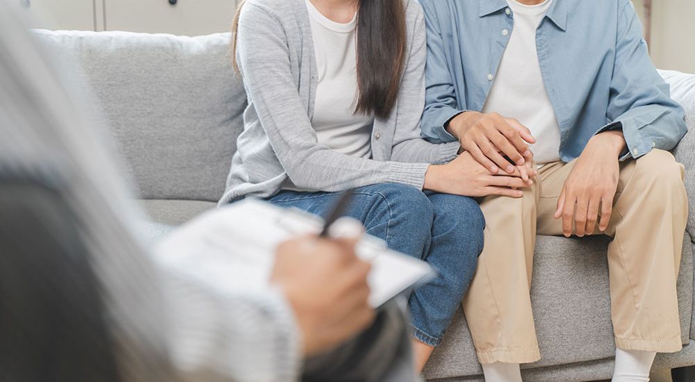 Couple holding hands during a therapy session on a gray sofa with therapist taking notes in the foreground.