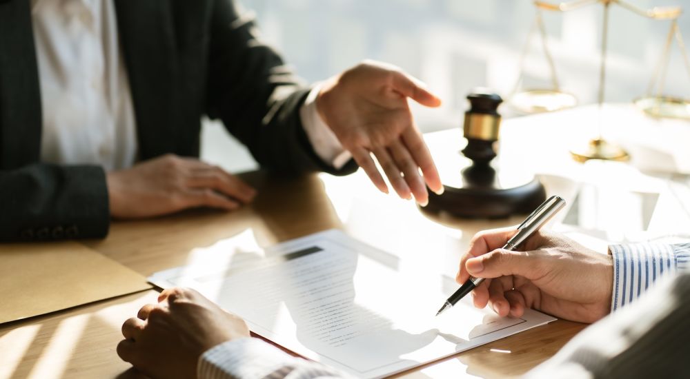 Two professionals discuss legal documents at an office desk with scales and a gavel in the background.
