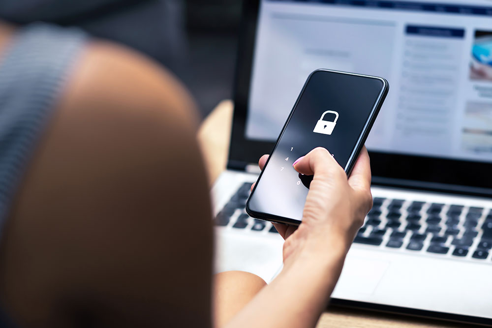 Person unlocking smartphone with security icon, seated at a laptop, illustrating cybersecurity and digital access.