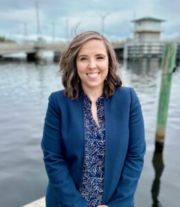 Smiling Woman in Blue Blazer Standing by Waterfront with Bridge and Cloudy Sky in the Background